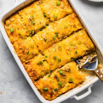 Sliced potato casserole in a white dish with a serving spoon, topped with melted cheese and chopped green onions, with one portion served on a plate.
