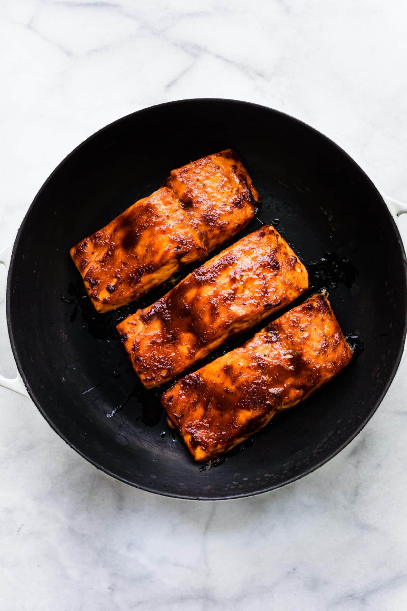 Overhead photo of three baked barbeque salmon filets in a cast iron skillet.