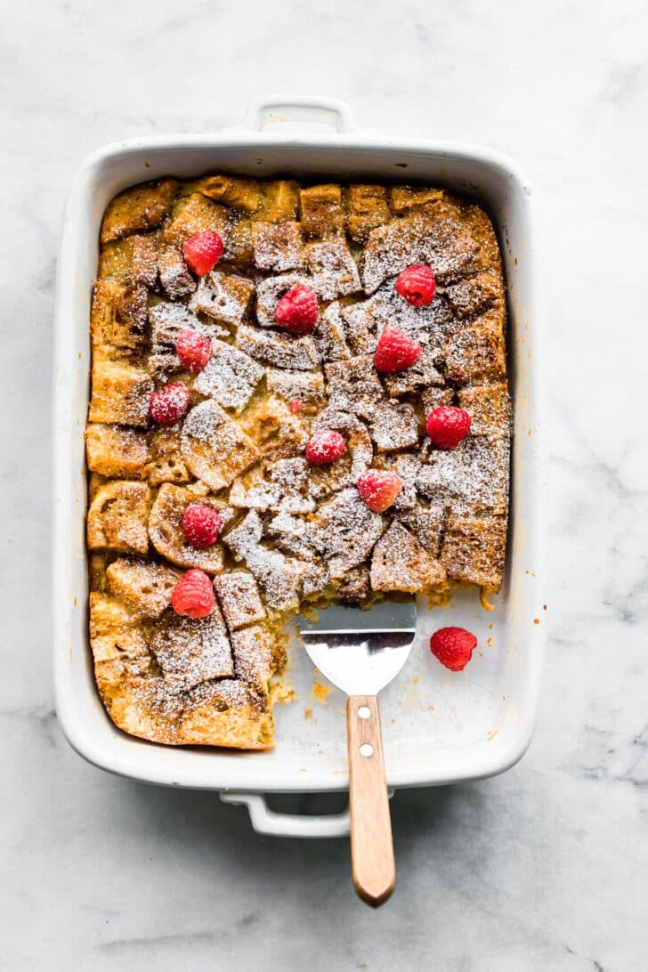 overhead photo of french toast casserole with raspberries in a white rectangle dish with a a serving utensil