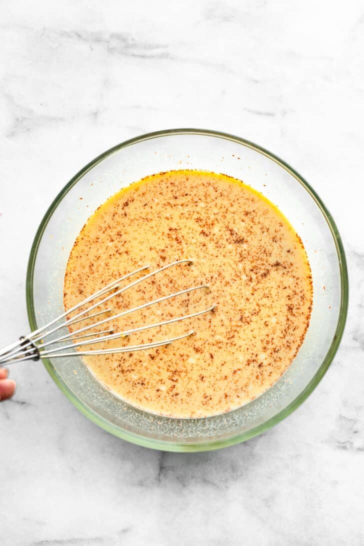 A whisk mixing together eggs, milk and spices in a glass mixing bowl.