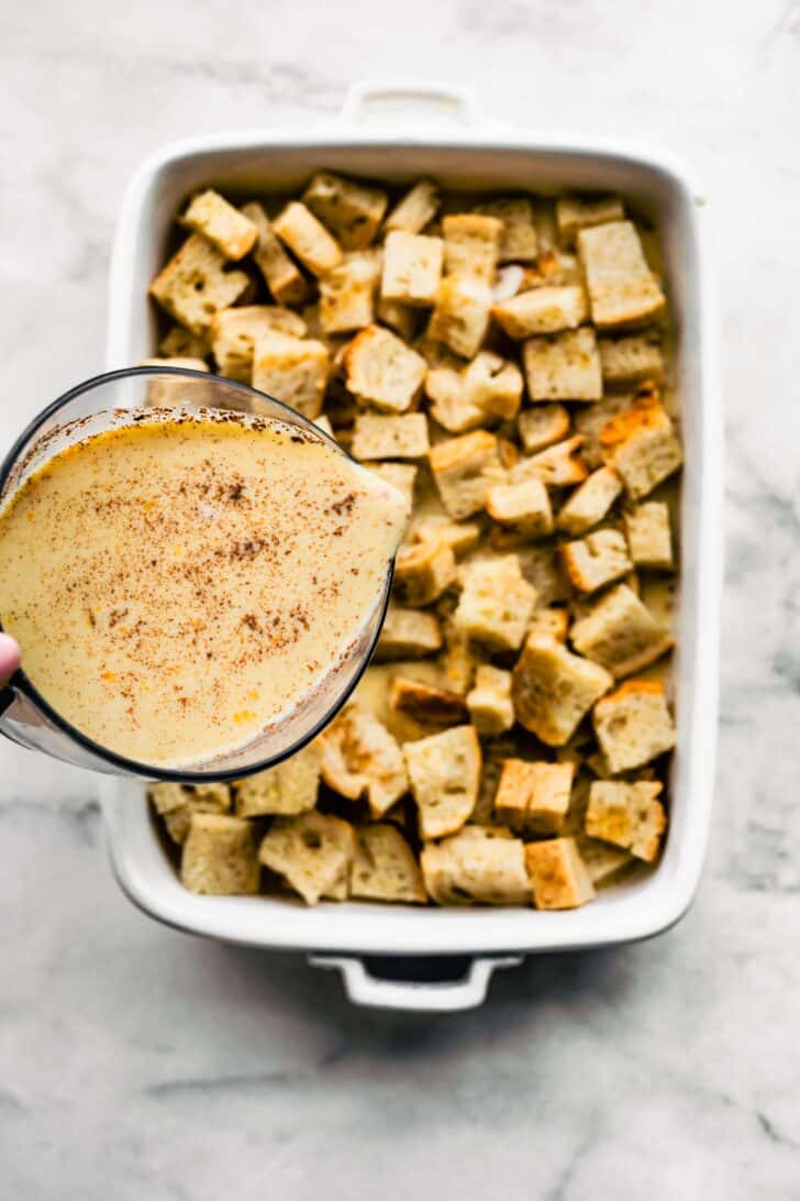 A custard mixture being poured over stale gluten free bread cubes in a baking dish.