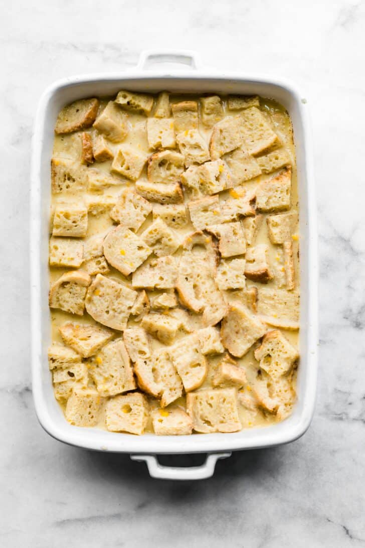 Stale bread soaking in an egg milk custard in a white 9x13 pan.