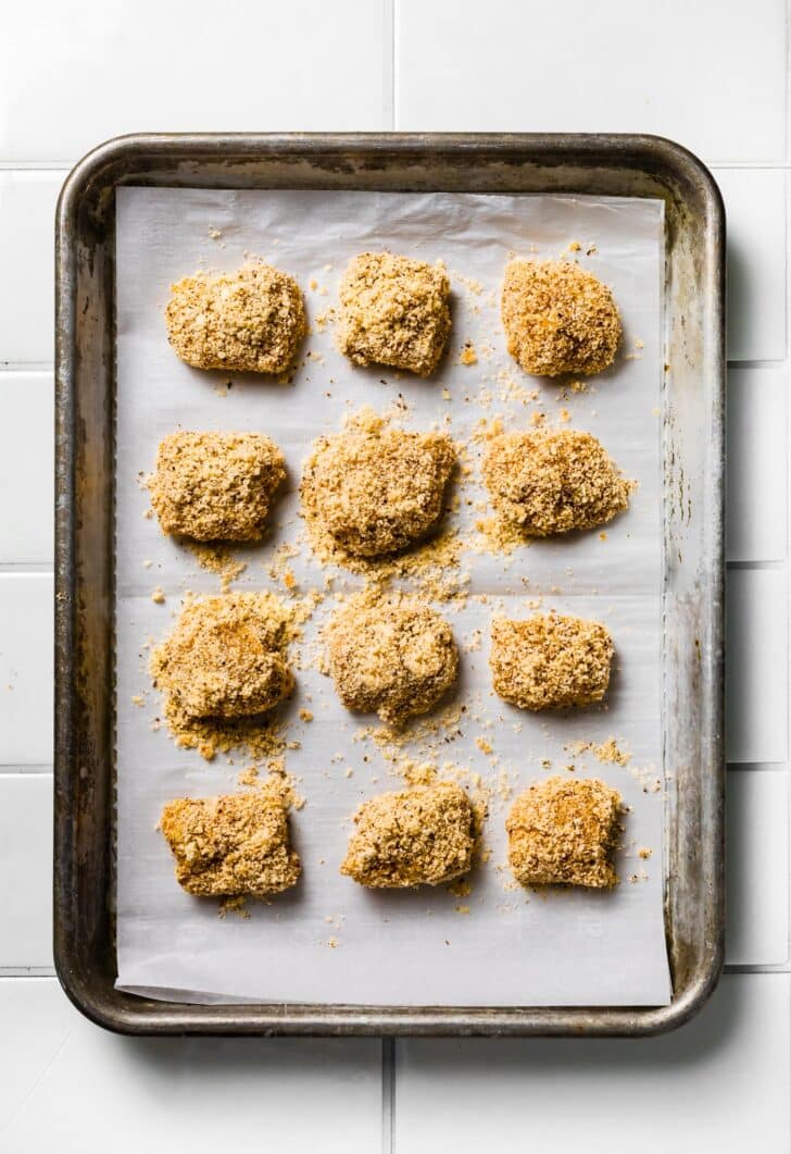 Uncooked breaded chicken nuggets arranged on a parchment-lined baking sheet before baking.