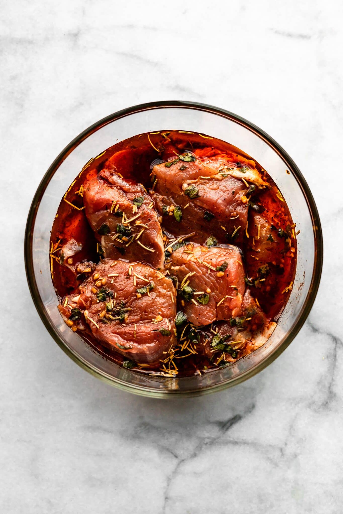 Pork medallions marinating in a bowl of herbs and sauces.