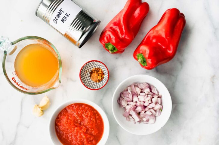 Red peppers, red onions, coconut milk, crushed tomatoes and broth scattered on a kitchen counter.