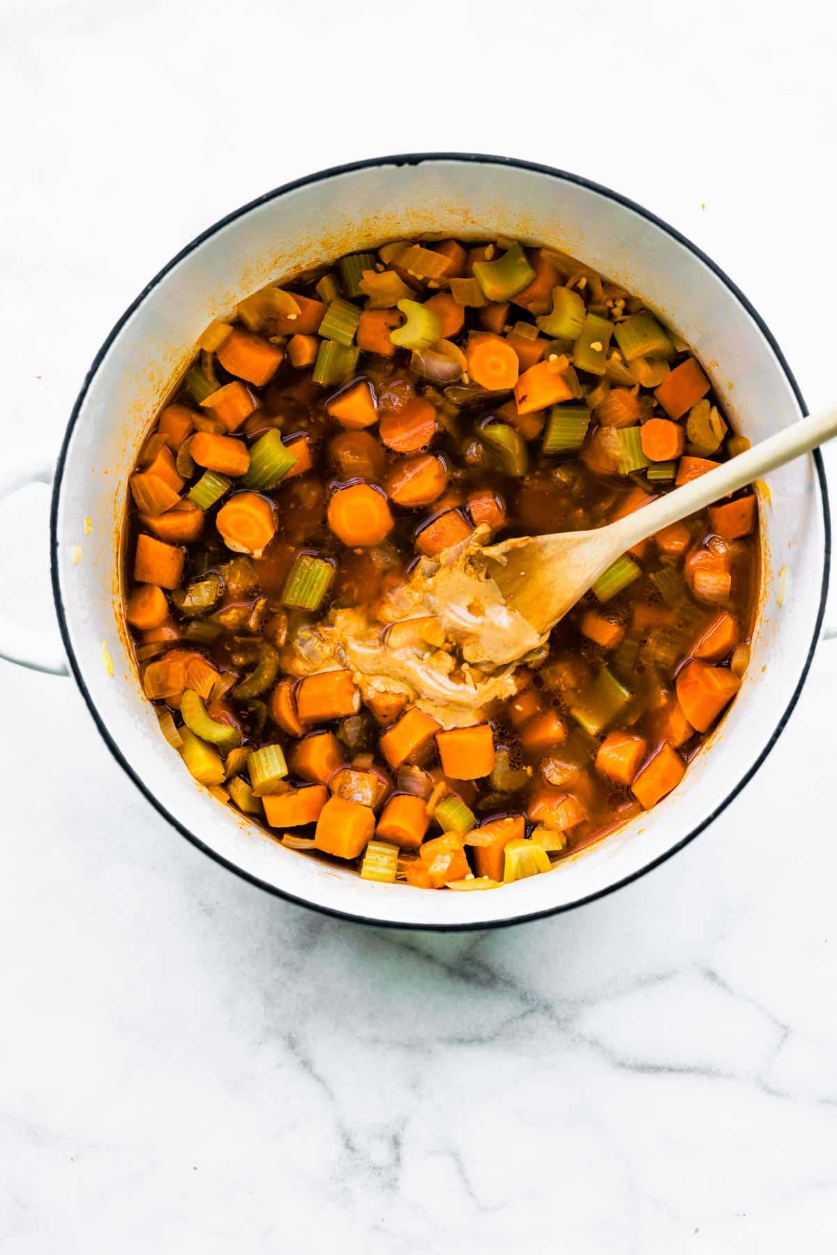 A wooden spoon full of peanut butter in a soup pot with broth and vegetables.