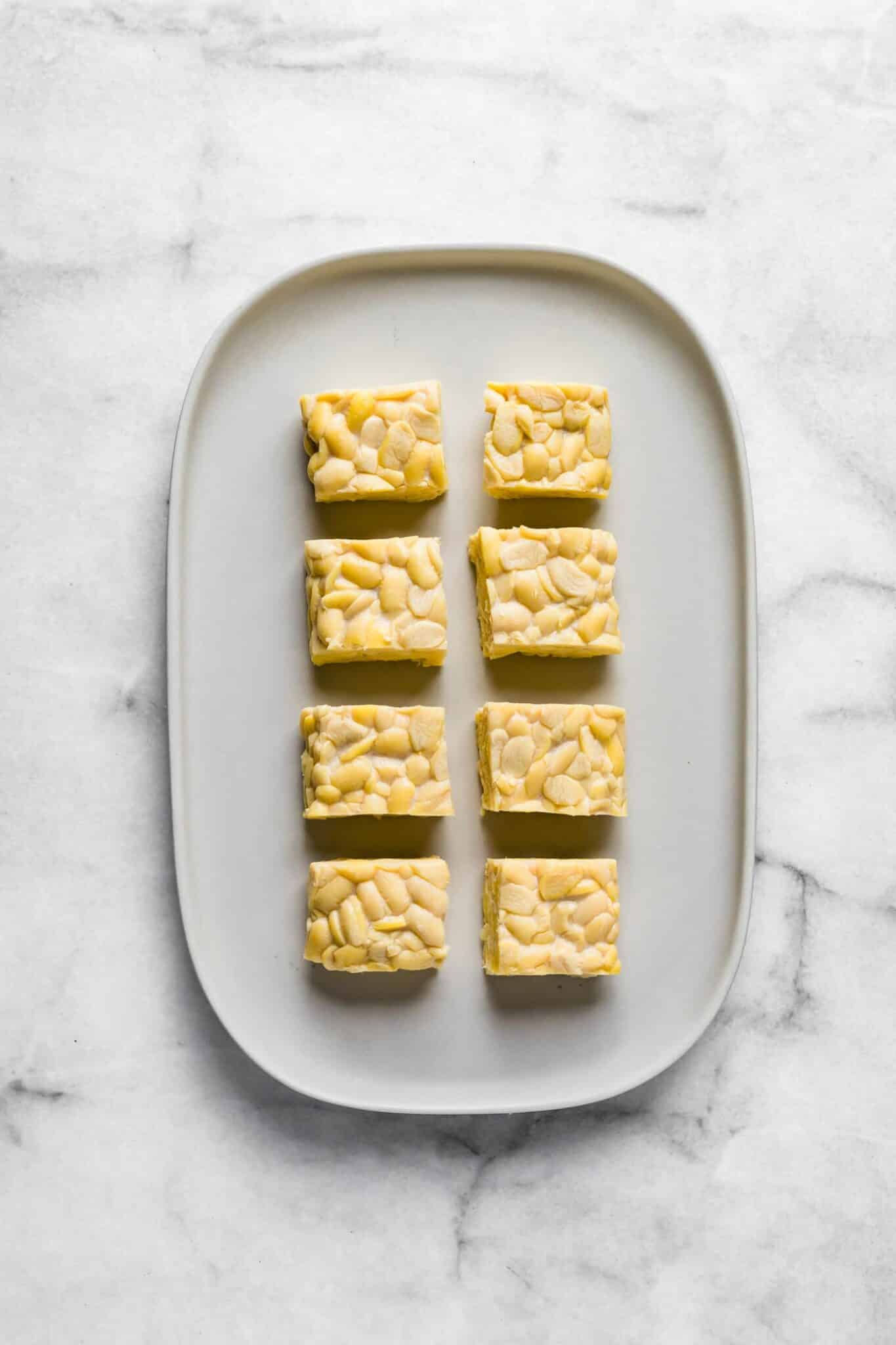 Eight cubes of raw tempeh on a white platter.