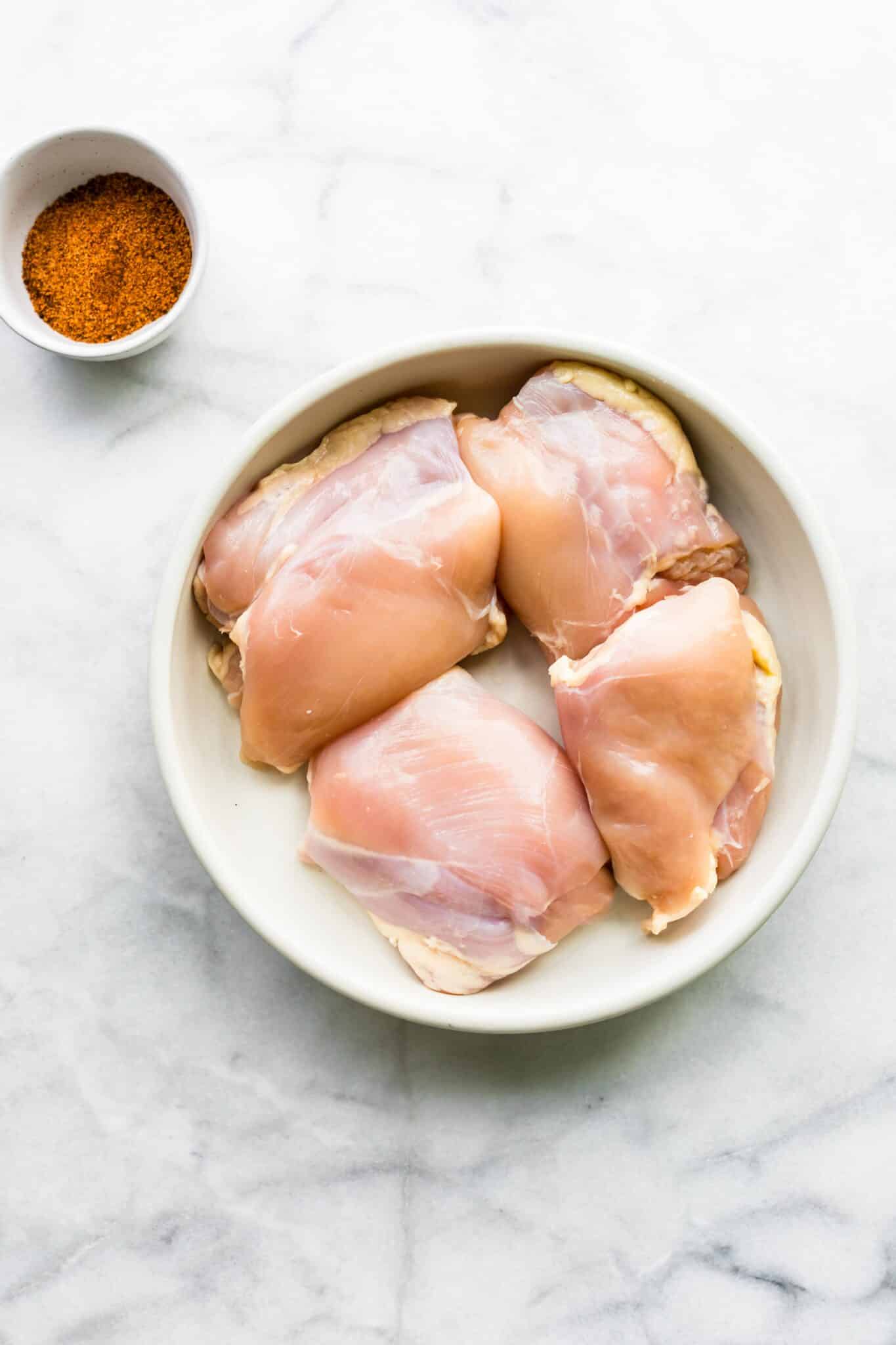 Overhead photo of raw chicken thighs in a bowl with a separate bowl of seasonings.
