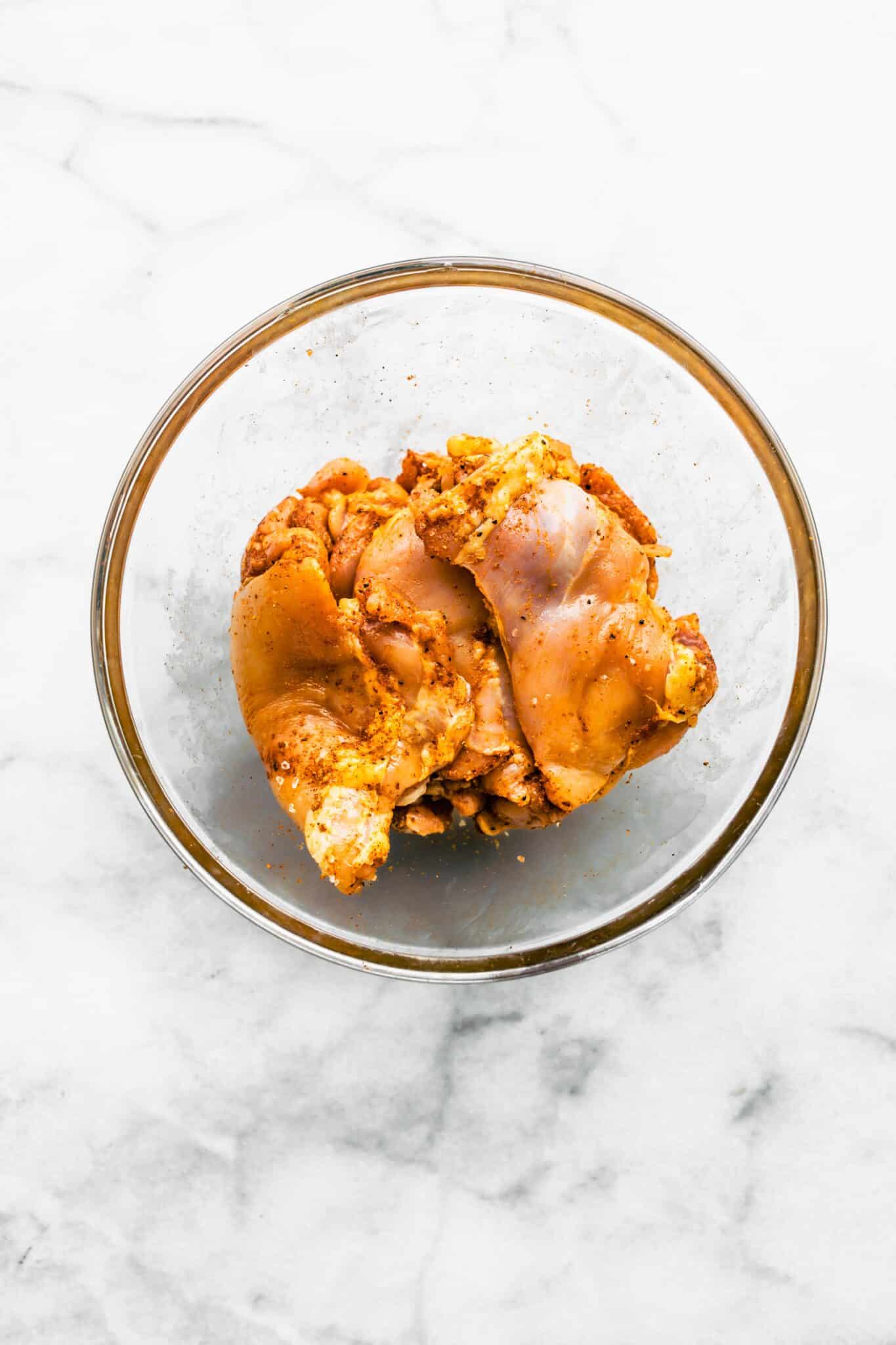 Overhead photo of seasoned raw boneless skinless chicken thighs in a glass bowl.