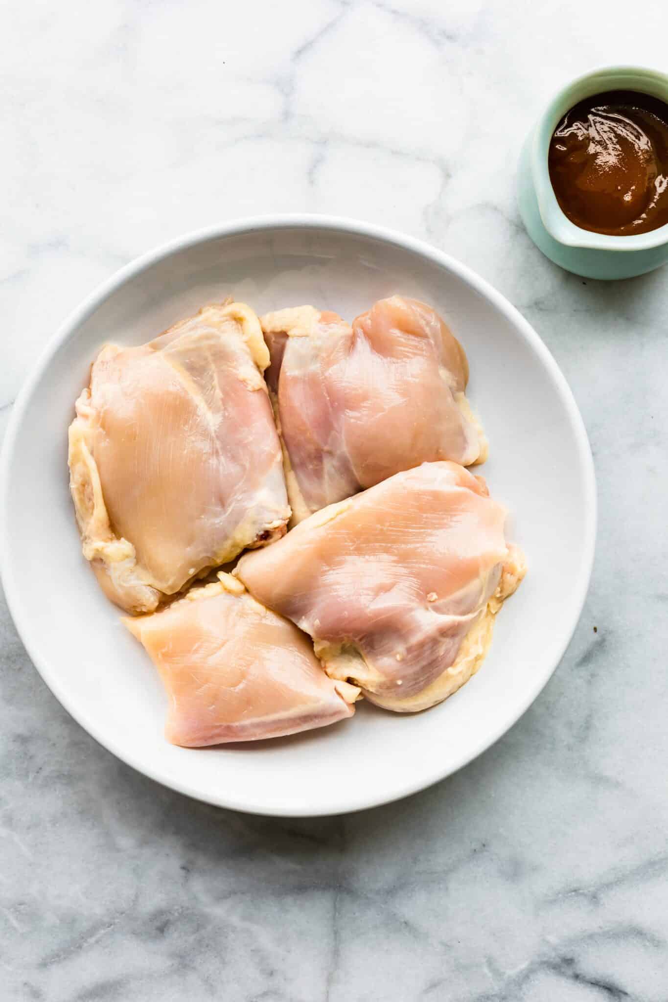Four chicken thighs on a white plate with BBQ sauce on the side.