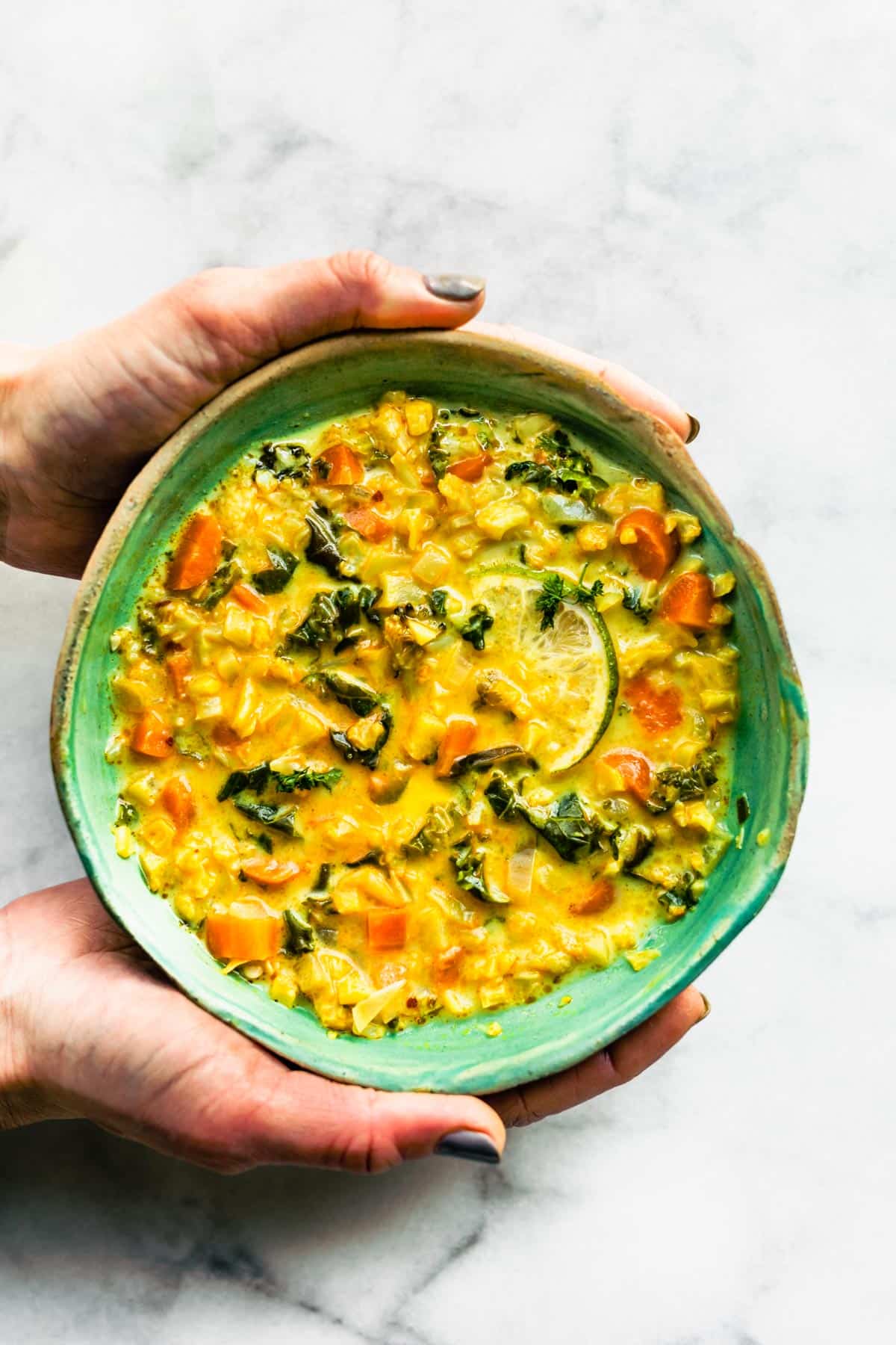 Hands holding a bowl of Curried Cauliflower Soup with Kale.