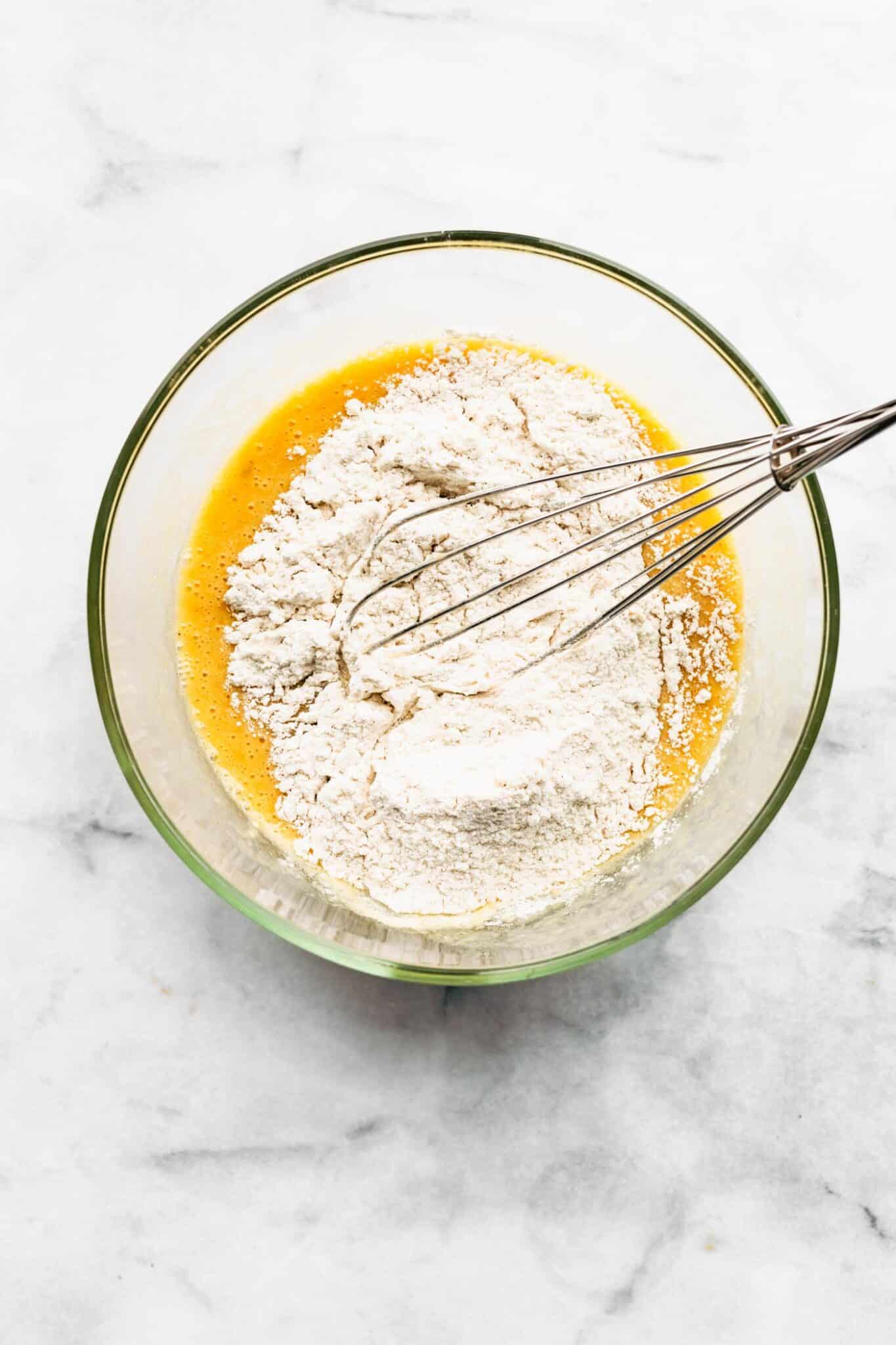 Gluten free flour and metal whisk on top of wet ingredients in a glass bowl.