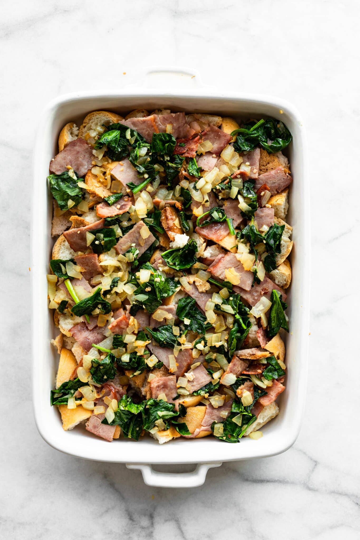 Overhead photo of a baking pan layered with gluten free bread, ham and sauteed veggies.