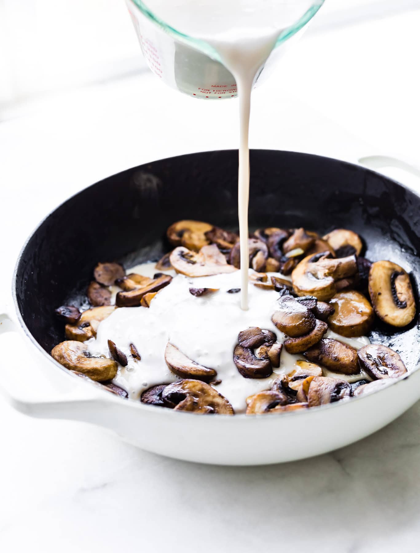 Coconut milk being poured into a pan of sauteed sliced mushrooms in a skillet.