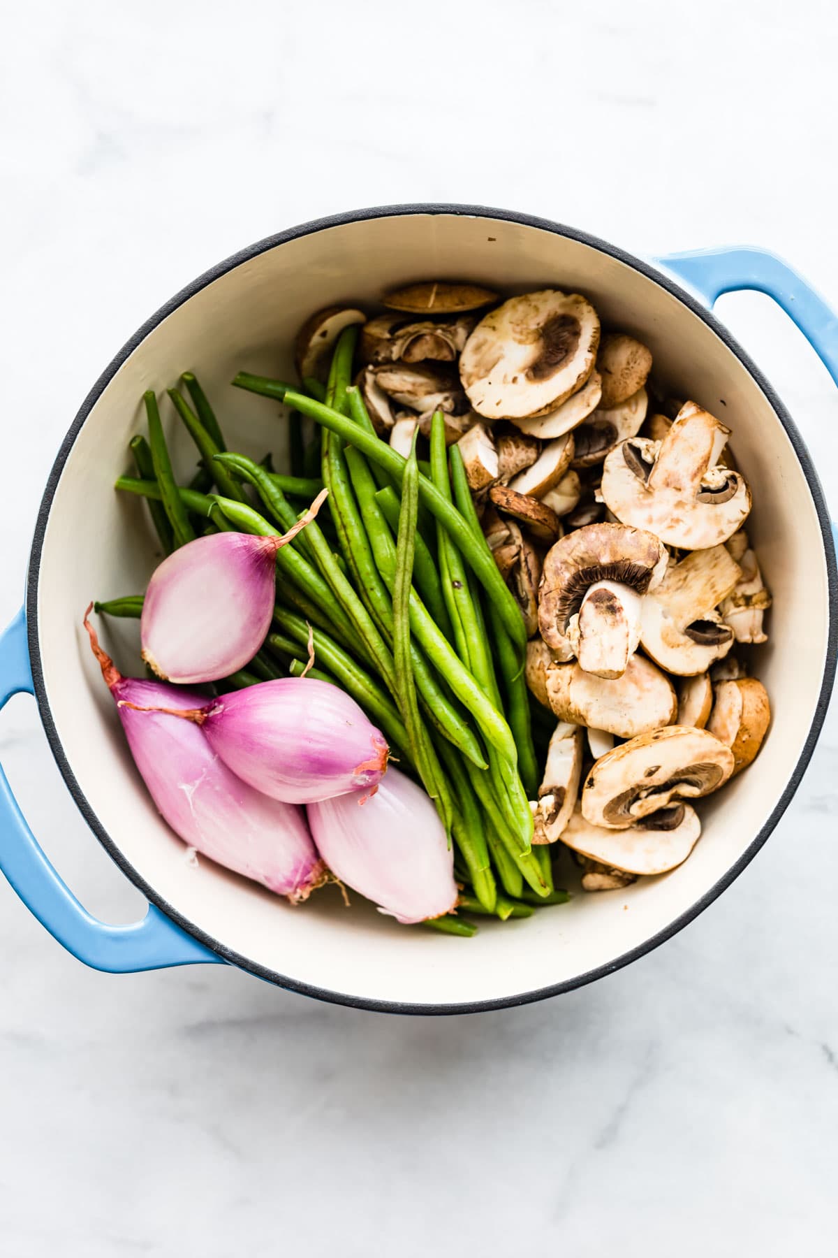 Fresh shallots, green beans, and mushrooms in a large white pot on a marble countertop.
