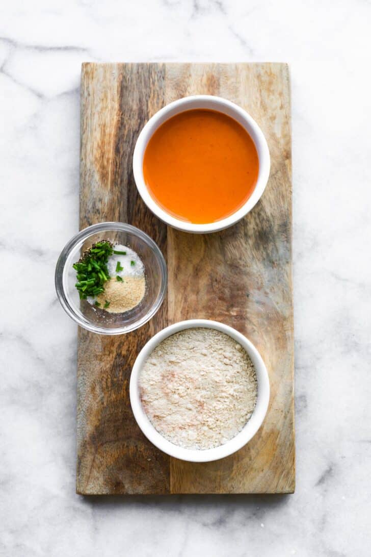 Buffalo sauce, seasonings, and flour in bowls on a wooden plank.