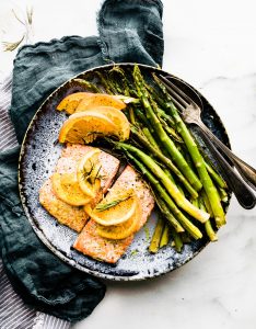 salmon on blue plate with orange slices on top and herbs. blue linen on the side. Fork on plate to the right of salmon. overhead angles shot