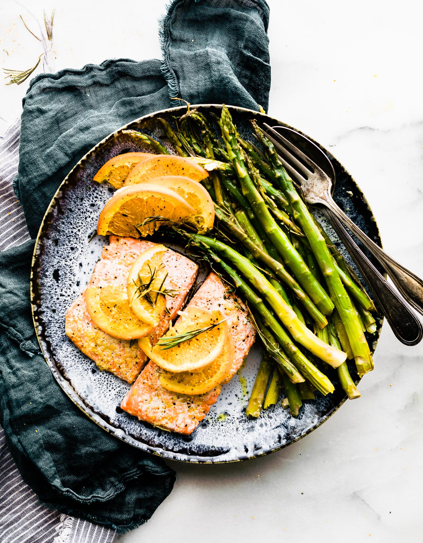 salmon on blue plate with orange slices on top and herbs. blue linen on the side. Fork on plate to the right of salmon. overhead angles shot