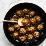 A saucy Asian meatball on a spoon over the pan of meatballs.