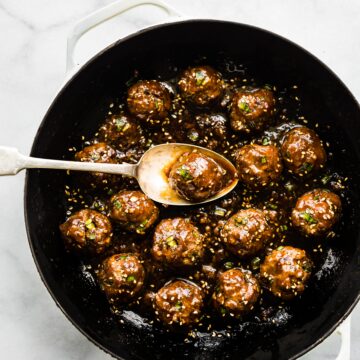 A saucy Asian meatball on a spoon over the pan of meatballs.