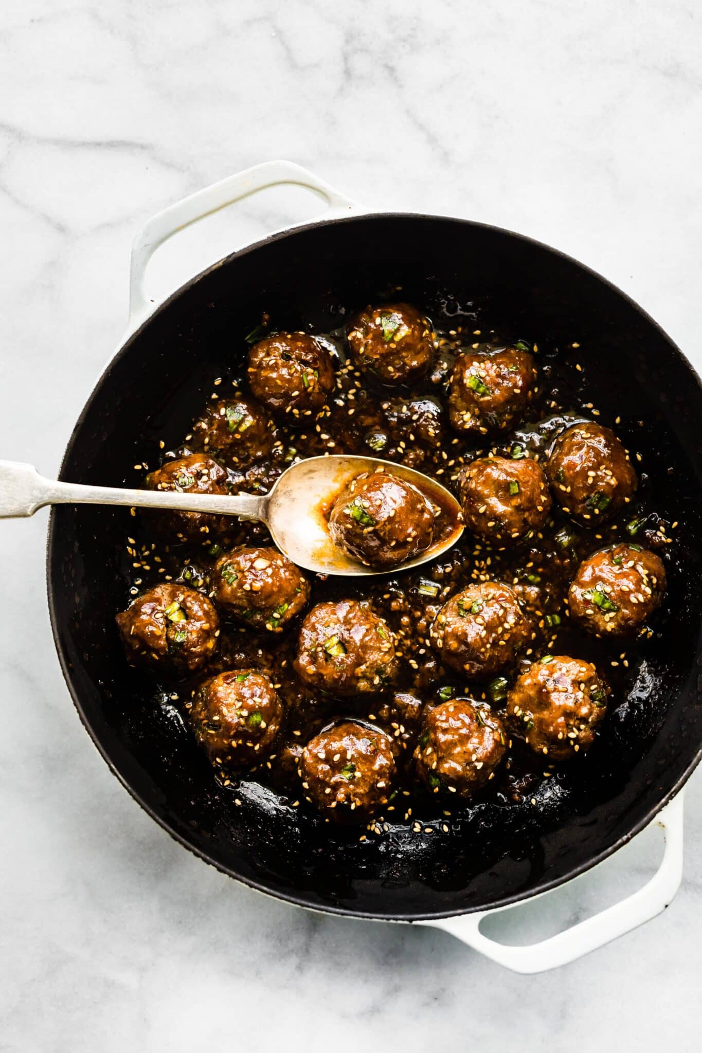 A saucy Asian meatball on a spoon over the pan of meatballs.