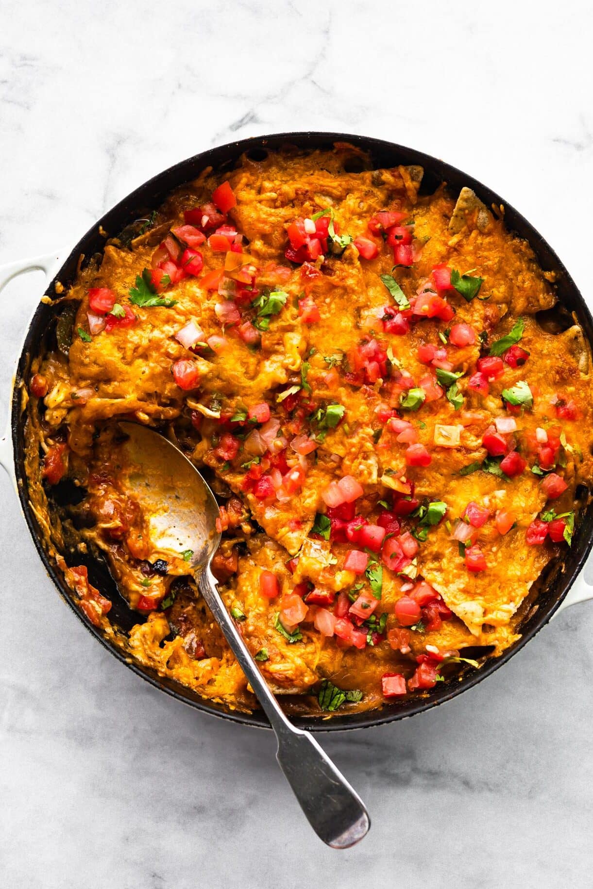 Chicken fajita casserole in a skillet after serving, showing melted cheese, tomato topping, and cilantro with a large spoon scooping out a portion.
