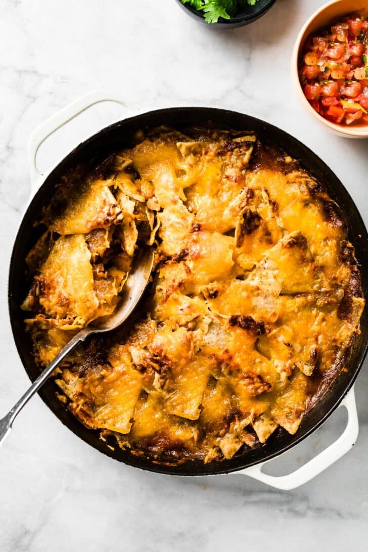 Baked chicken fajita casserole in a skillet with a serving spoon, no toppings. Small side bowls of fresh tomato and cilantro sit nearby.