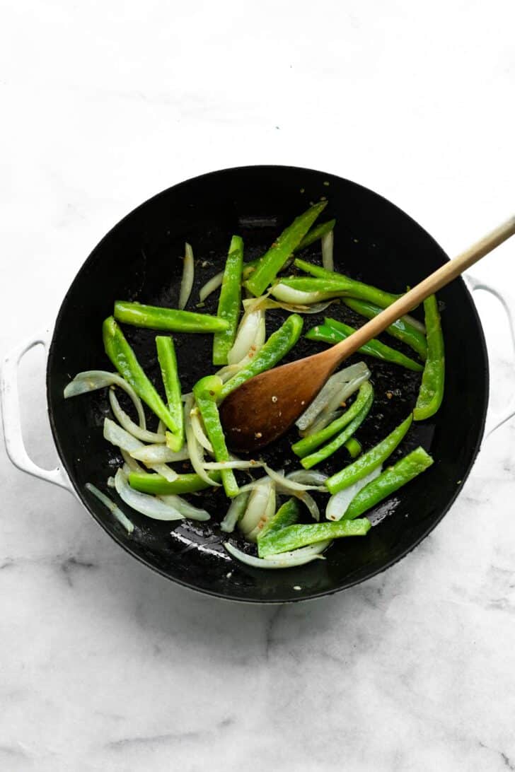 Freshly sautéed green bell peppers and onions in a black skillet with a wooden spoon.