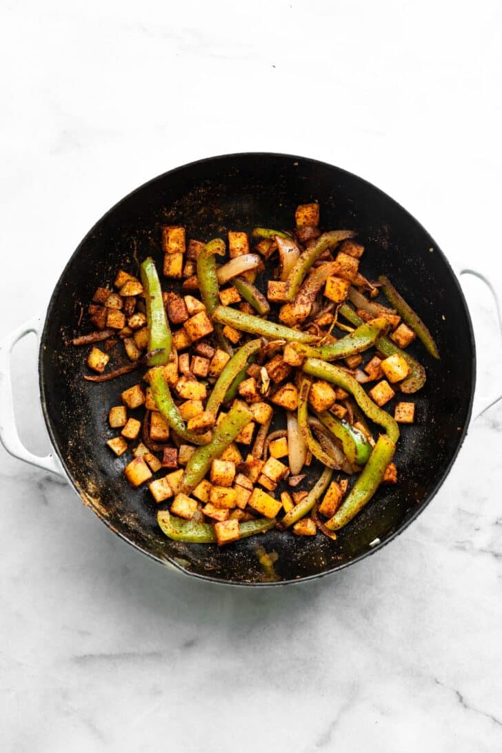 seasoned Diced potatoes, green bell peppers, and onions sautéing in a black skillet with a wooden spoon.