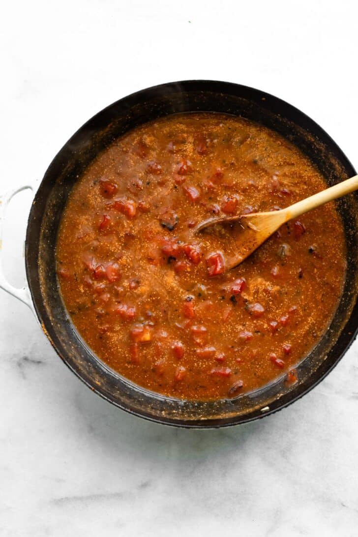 Tomato-based sauce with spices simmering in a skillet, stirred with a wooden spoon.