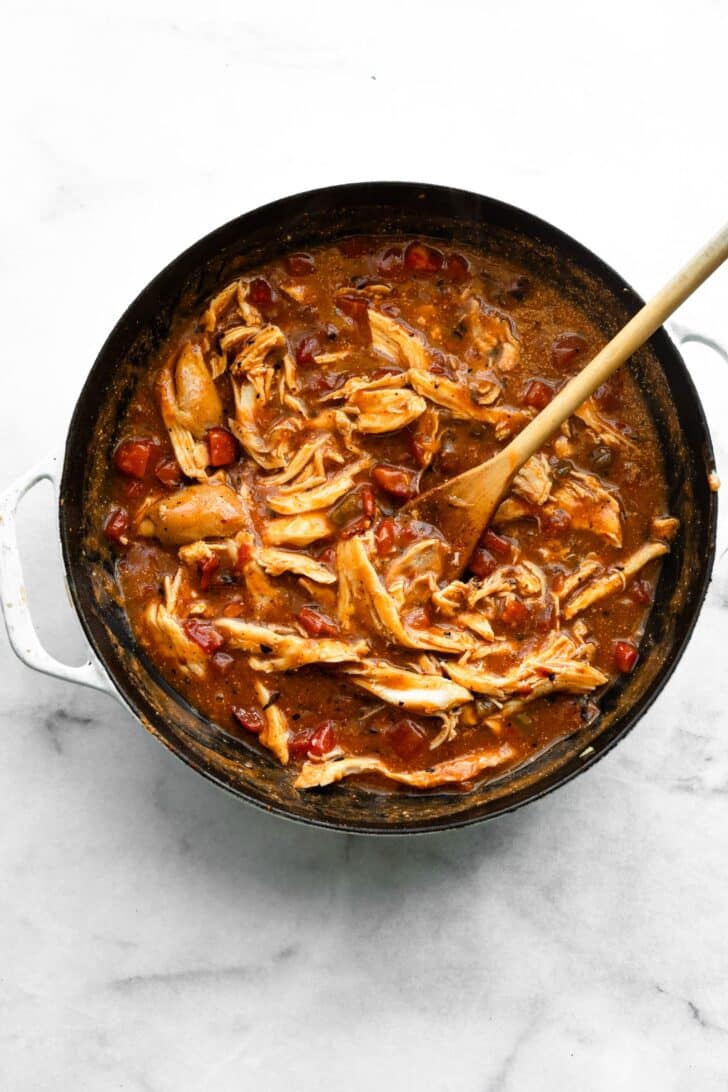 Shredded chicken mixed into a tomato-based sauce in a skillet, stirred with a wooden spoon.