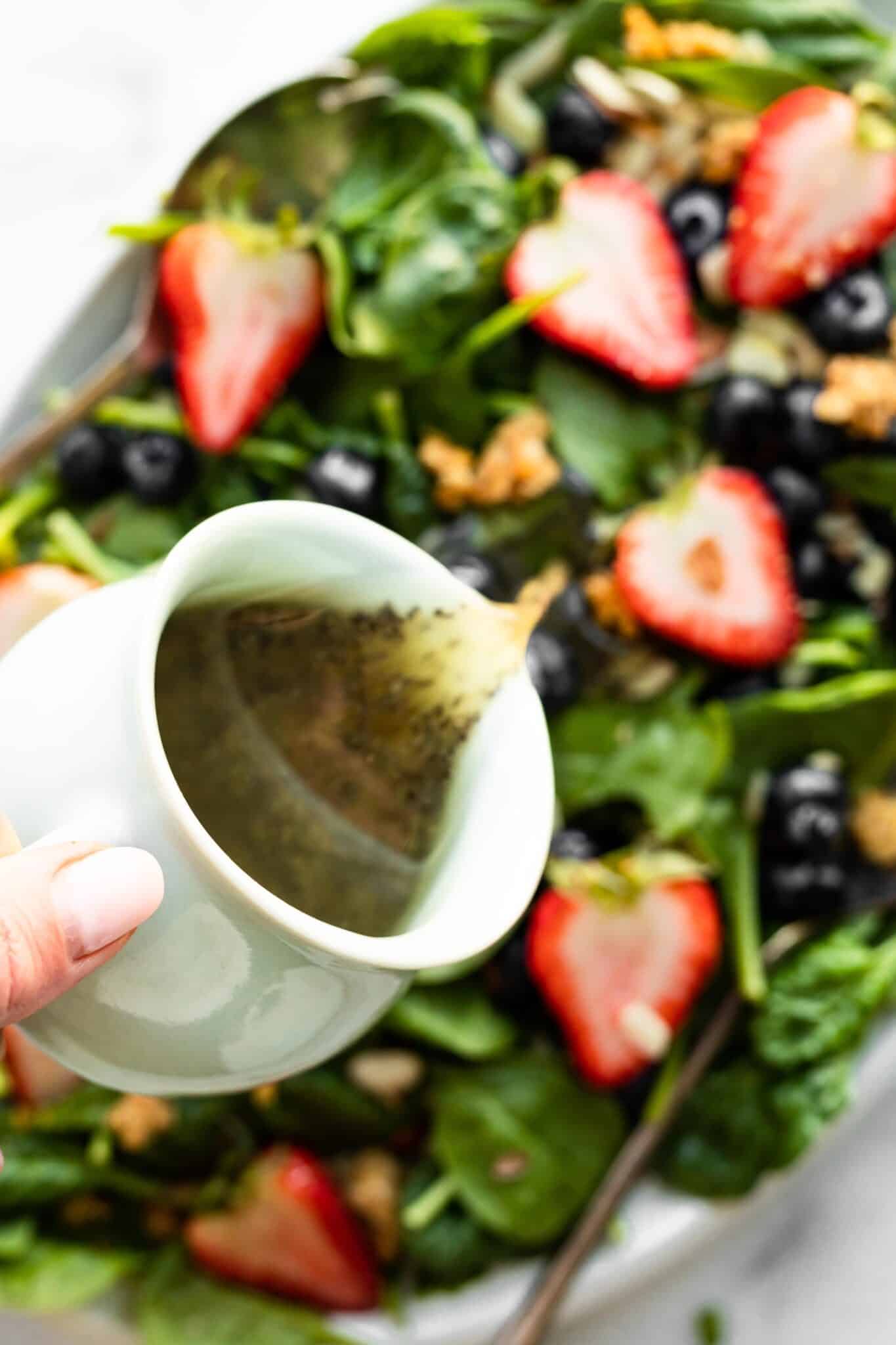 A woman's hand pouring poppyseed dressing from a small white carafe onto a berry salad.