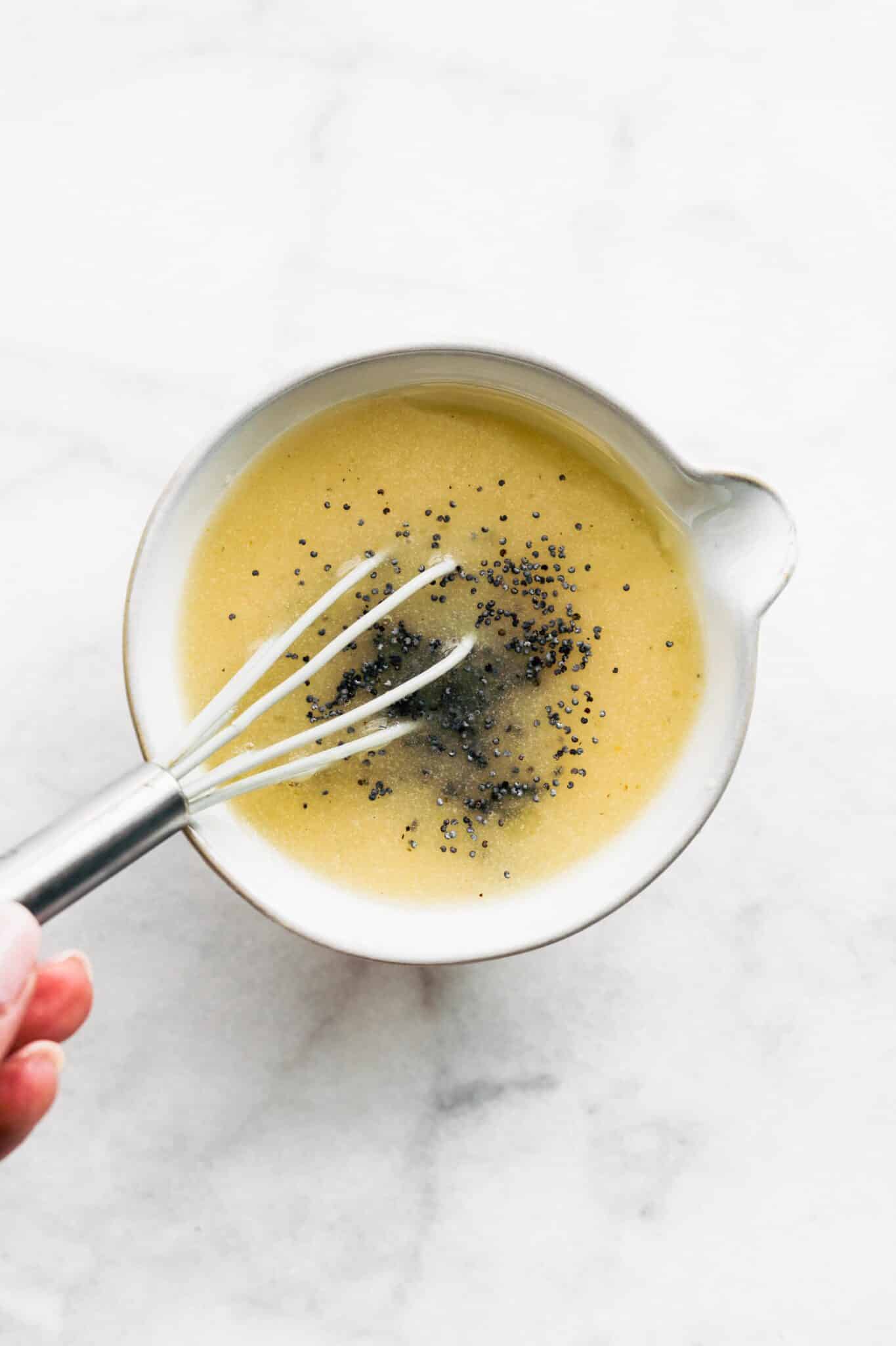 A small whisk whisking poppy seeds into a salad dressing in a white bowl.