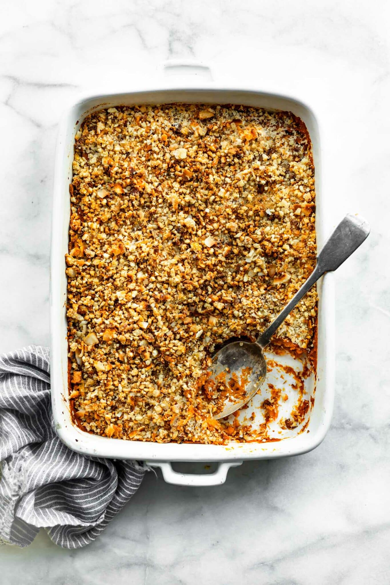 White casserole dish filled with vegan sweet potato casserole with a corner missing and a serving spoon