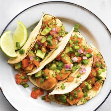 3 shrimp tacos on a white plate topped with avocado and chopped onion and a lime wedge on the plate