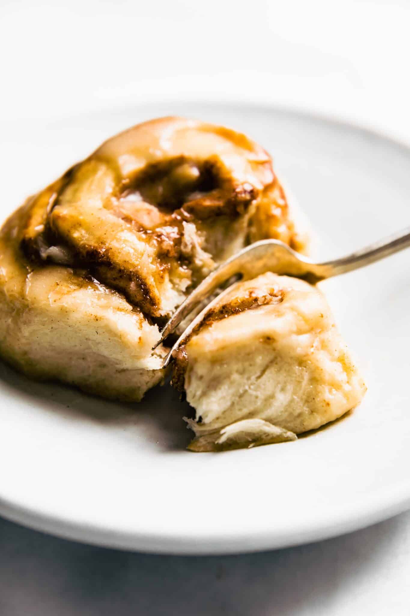 gluten free cinnamon roll on white plate with a fork cutting through it. Side angle