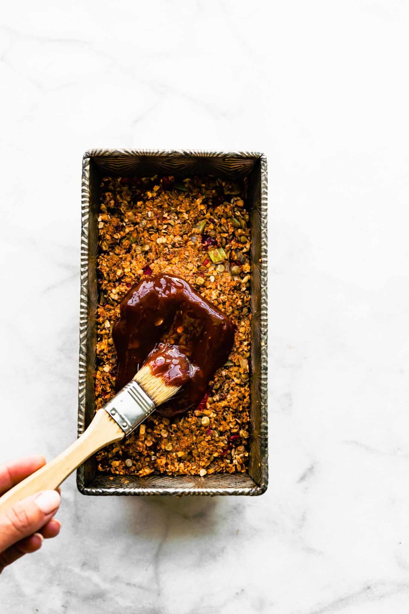 A woman's hand using a brush to spread a ketchup sauce on a lentil loaf.