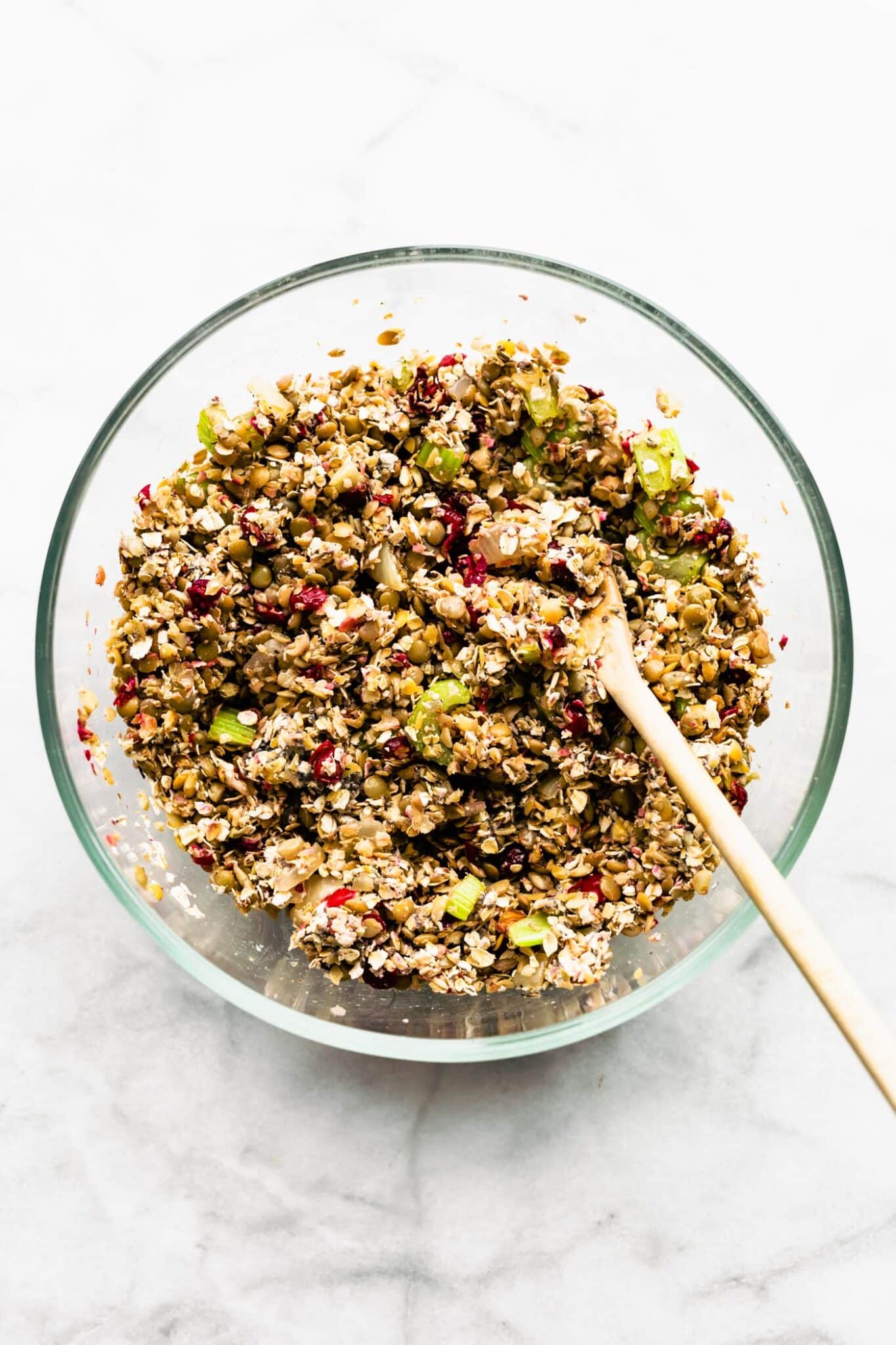 A wooden spoon mixing lentils, sauteed veggies and cranberries in a glass mixing bowl.