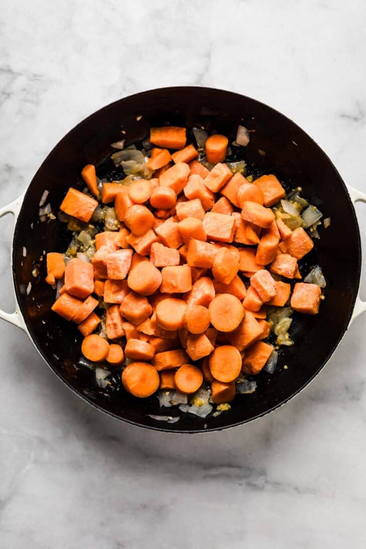 Diced carrots and sweet potatoes added to sautéed onions and garlic in a black skillet, creating the vegetable base for African peanut stew.