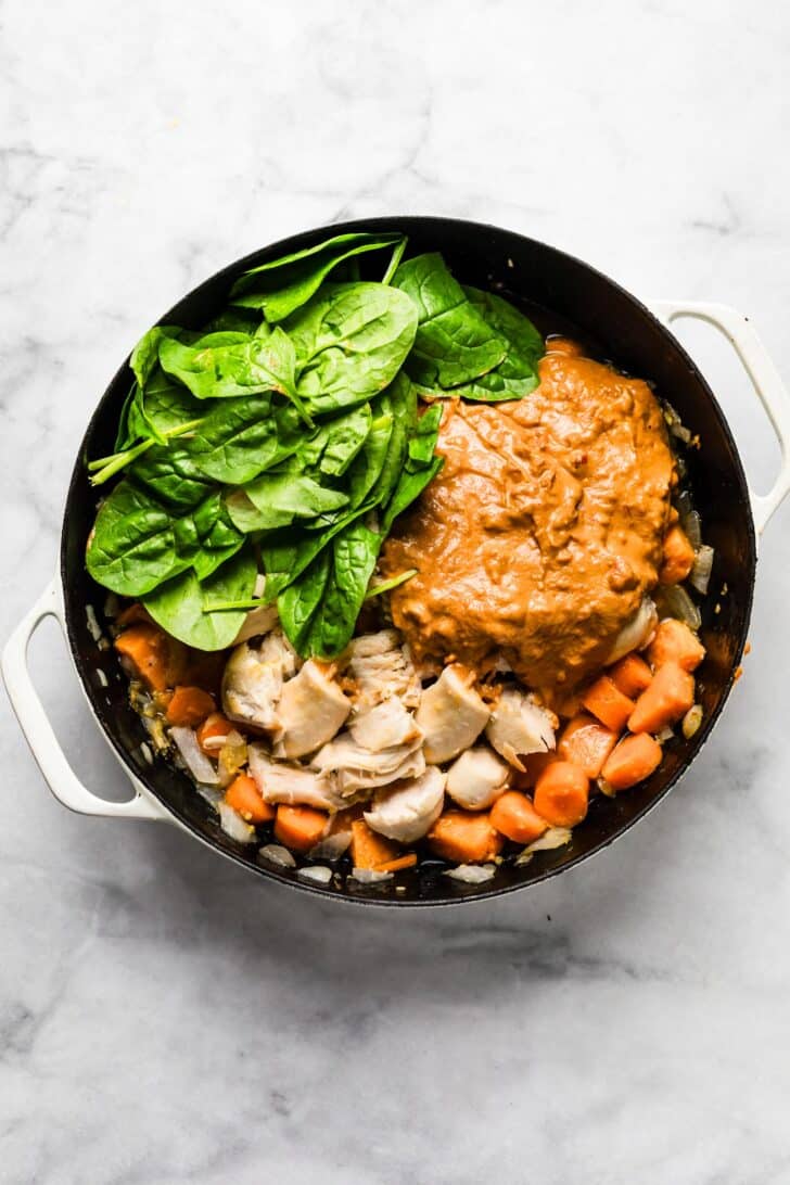 Large skillet filled with cooked chicken pieces, baby spinach leaves, and a thick peanut-tomato sauce mixture before simmering