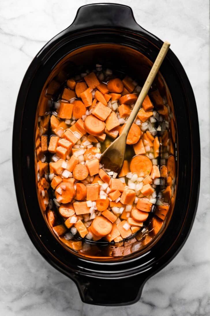 Cubed sweet potatoes, sliced carrots, and chopped onions in a slow cooker with a wooden spoon,