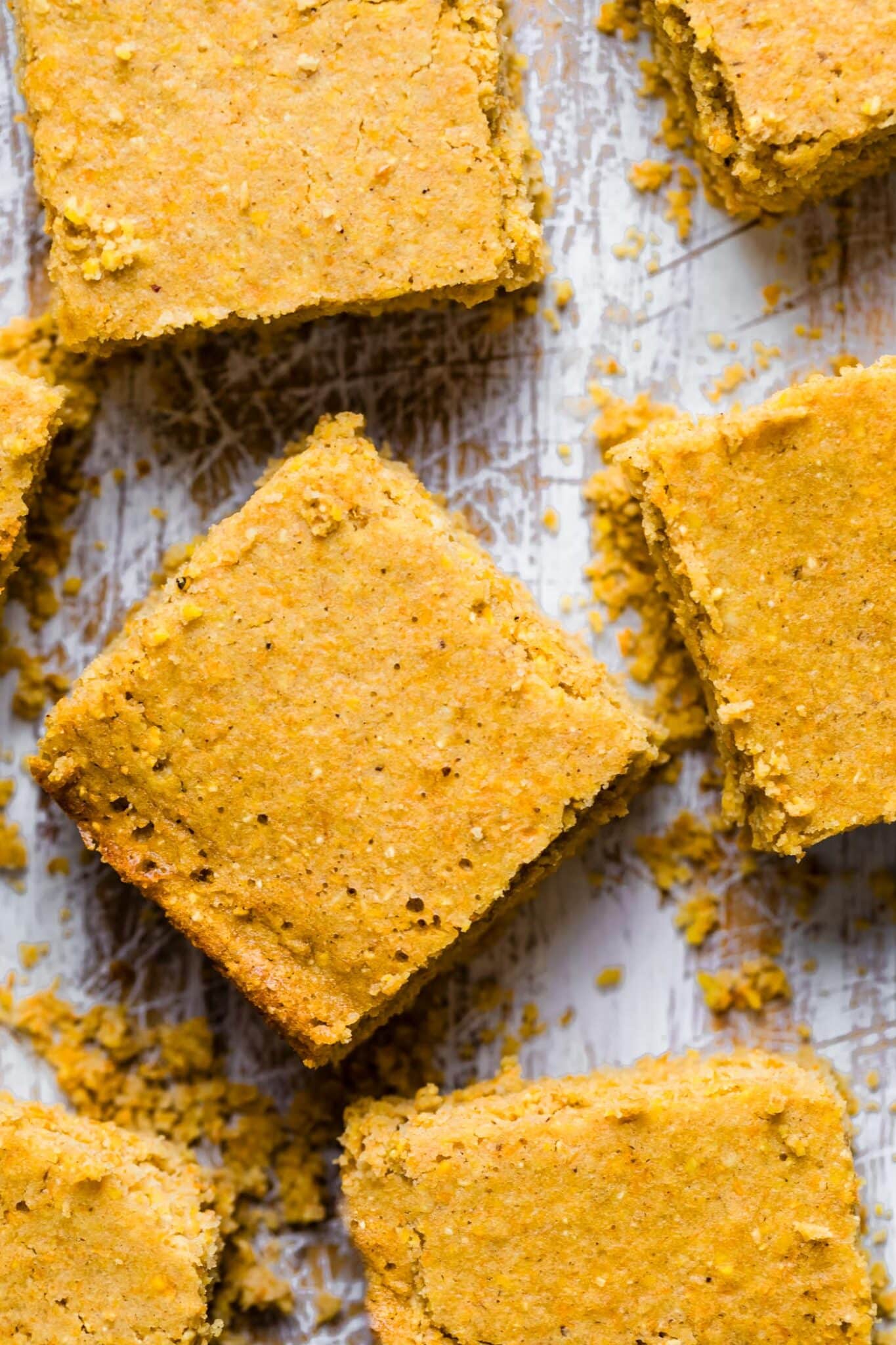 Close-up overhead shot of gluten-free cornbread squares arranged on a white wood surface with crumbs scattered around.