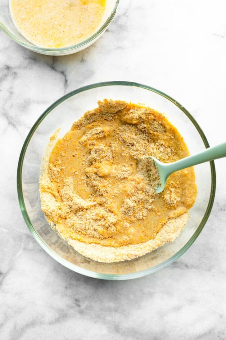 A glass mixing bowl with gluten-free cornbread batter being stirred with a spatula, showing the dry cornmeal mixture partially combined with the wet ingredients.