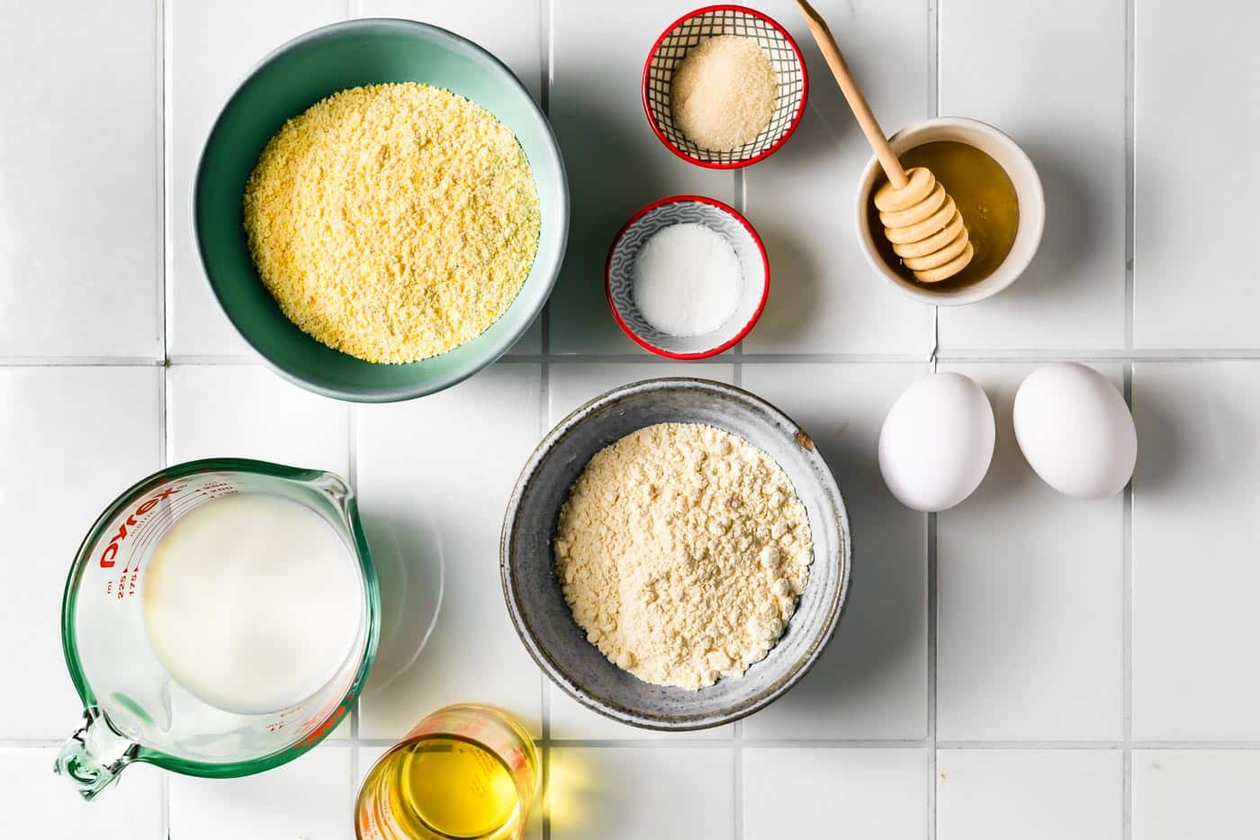 Flat lay of gluten-free cornbread ingredients, including cornmeal, flour, eggs, honey, salt, baking powder, milk, and oil arranged in bowls and measuring cups.