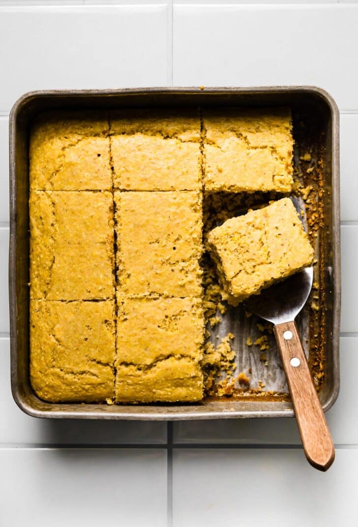 Baked gluten-free cornbread cut into squares in a metal pan, with one piece lifted on a serving spatula.