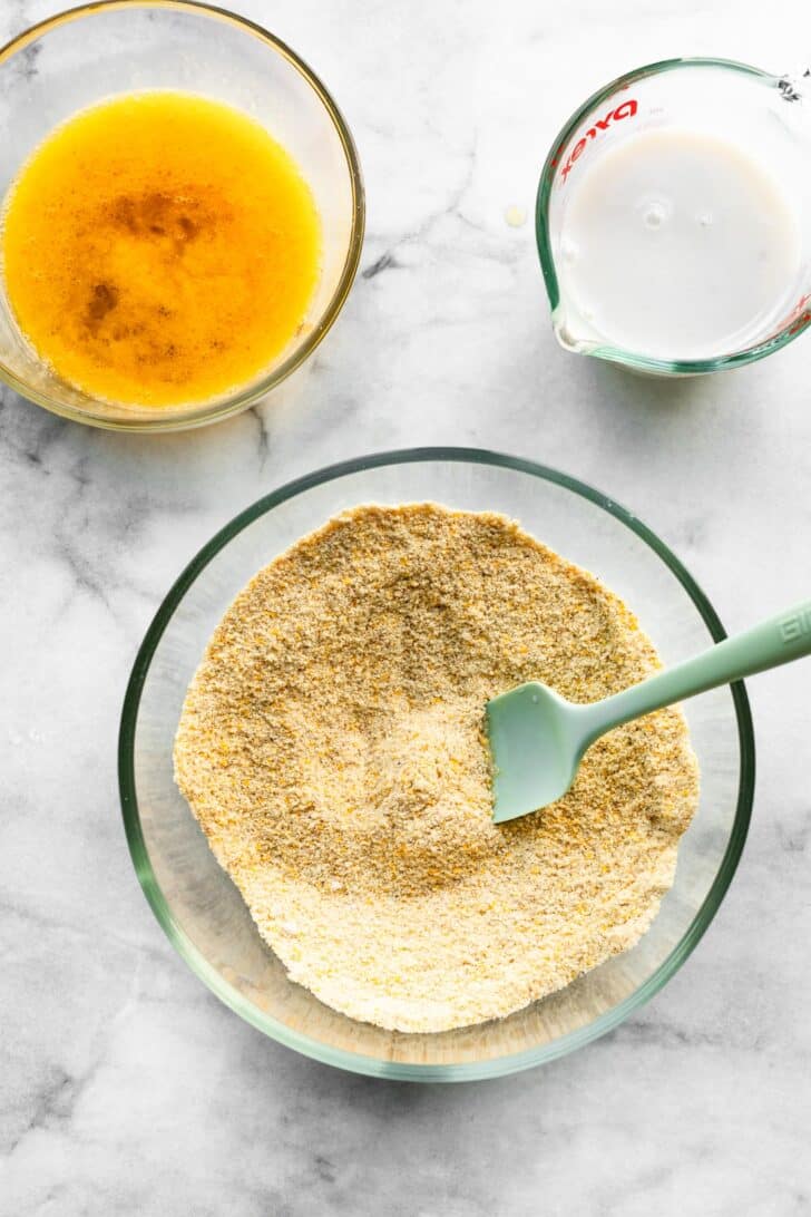 Dry ingredients for gluten-free cornbread in a mixing bowl with a spatula, next to bowls of melted butter and milk.