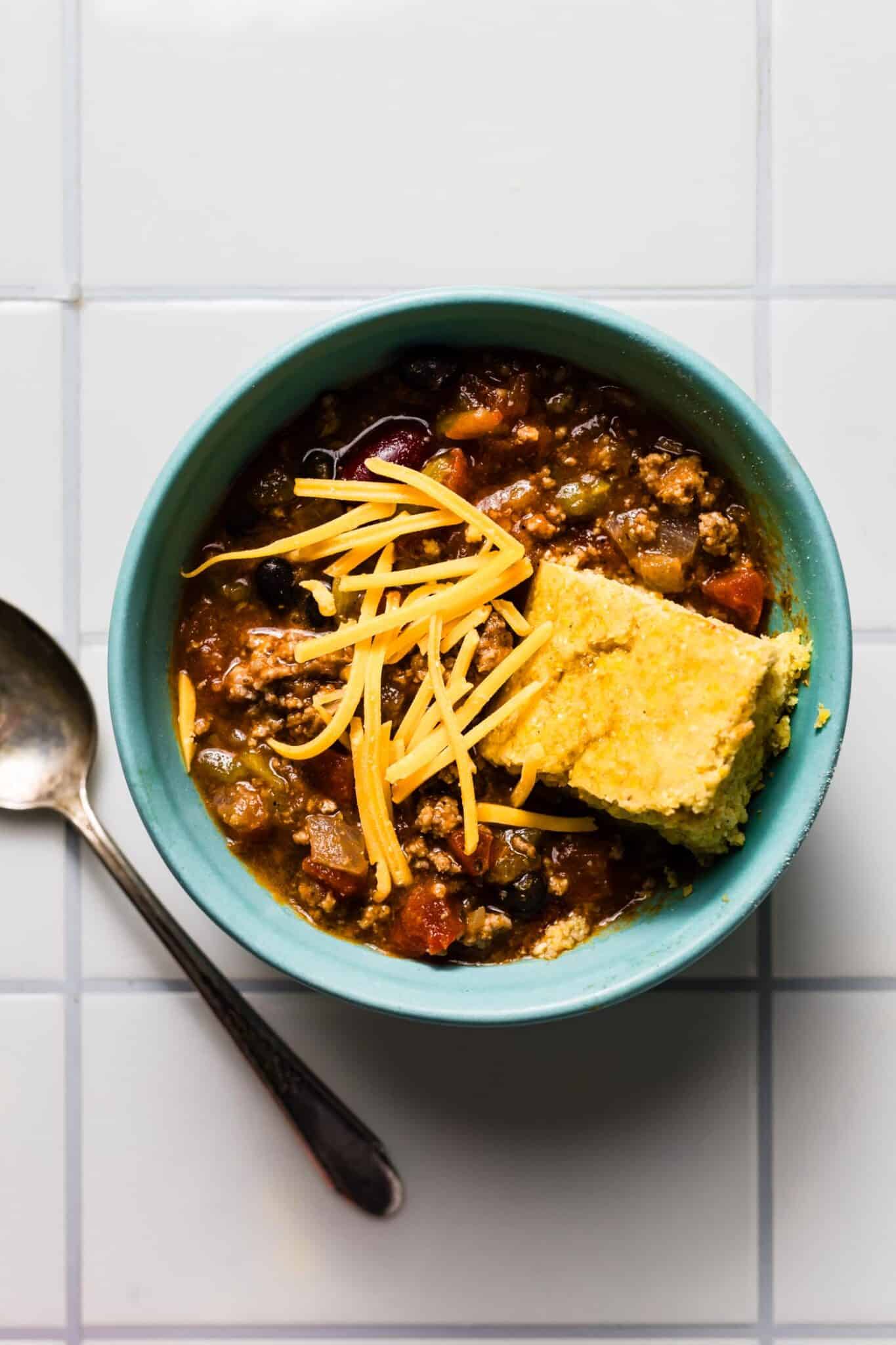 A bowl of chili topped with shredded cheddar cheese and a square of gluten-free cornbread resting in it