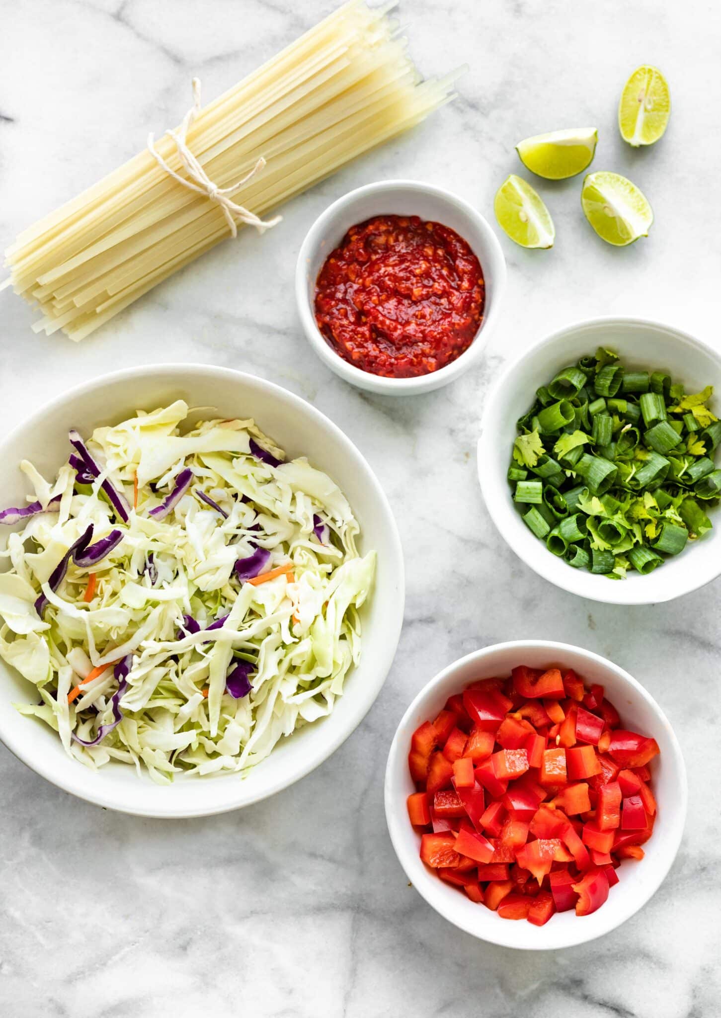 Photo of a cabbage mix, diced bell pepper, and uncooked rice noodles in separate bowls.