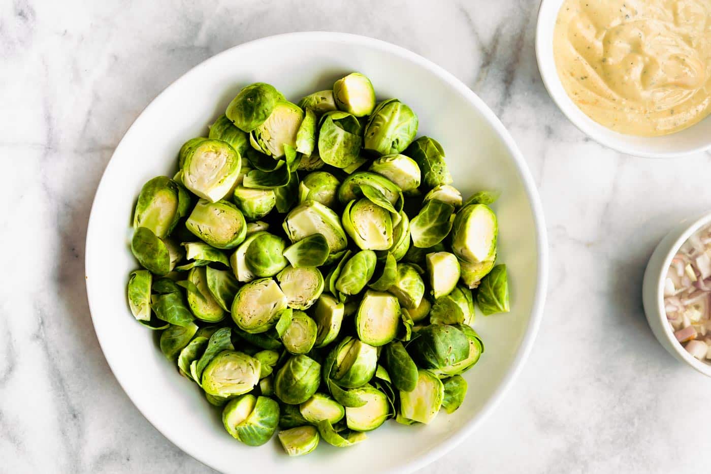 Halved brussels sprouts in a white bowl with bowls of dressing and diced onion.