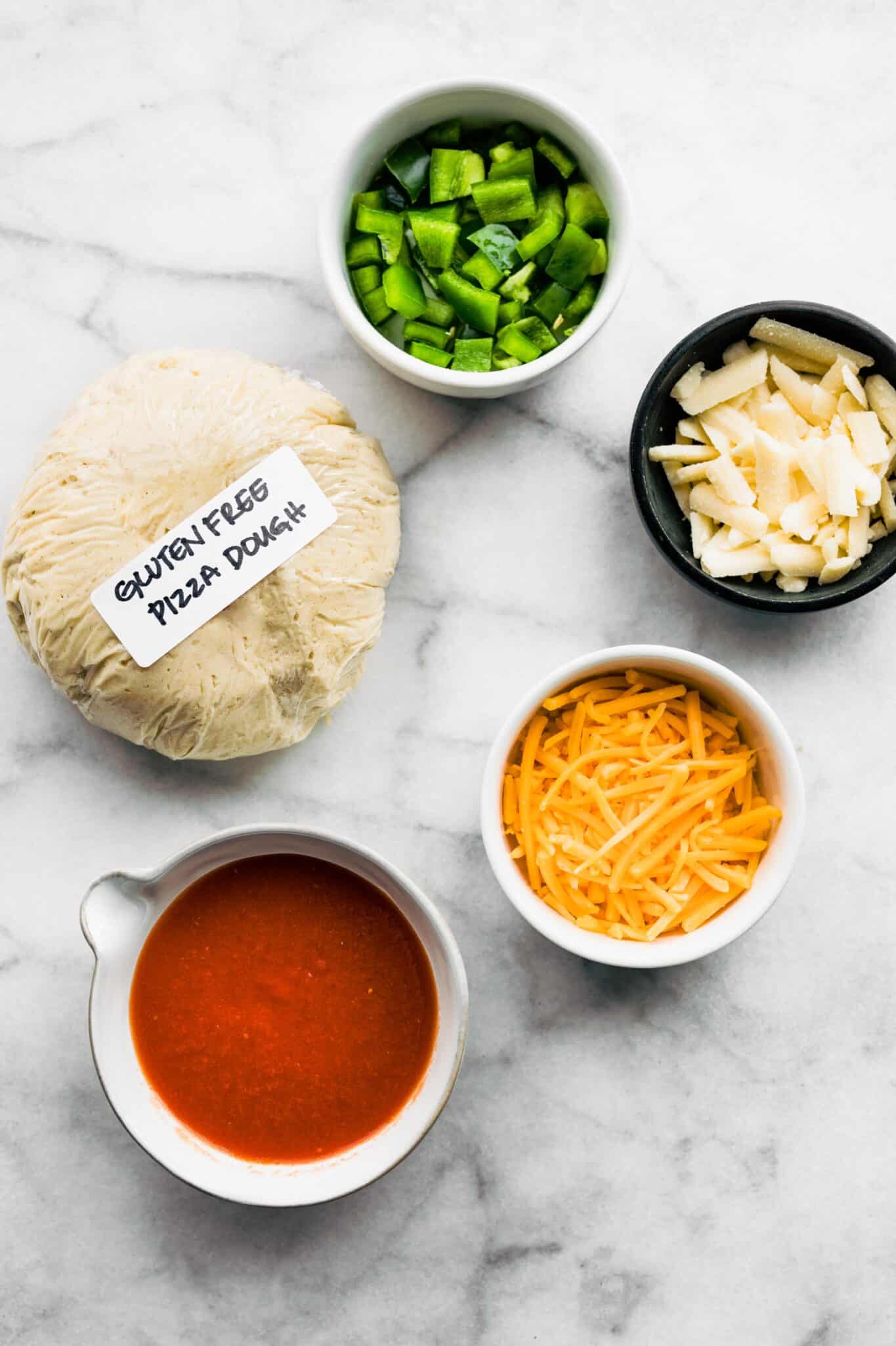 Overhead photo of a gluten free pizza dough puck, pizza sauce, cheese, and peppers.